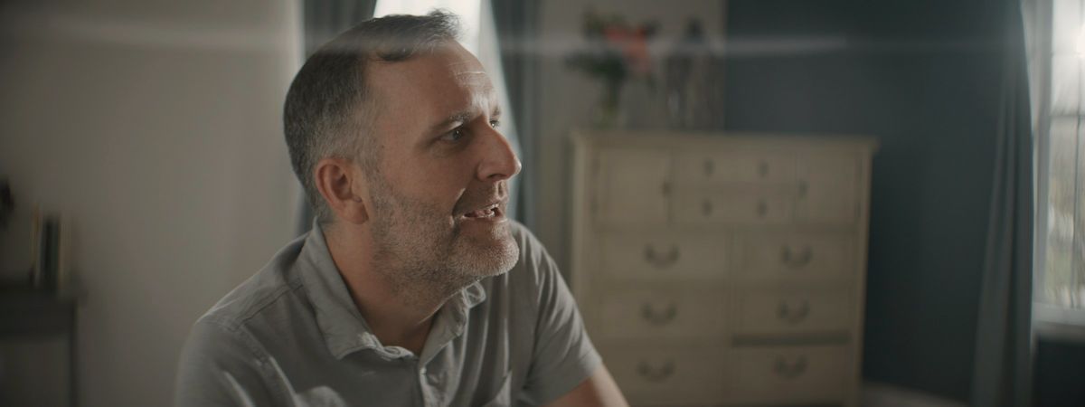 A man in his fifties talking, mid-sentence, in a warm, sunlit room